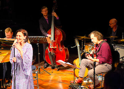 Das Bild zeigt eine Jazz-Band während eines Auftritts auf einer beleuchteten Bühne. Im Vordergrund sieht man eine Sängerin in einem lila Kleid mit Mikrofon, während im Hintergrund Musiker mit Kontrabass, Gitarre, Schlagzeug und Klavier zu sehen sind.