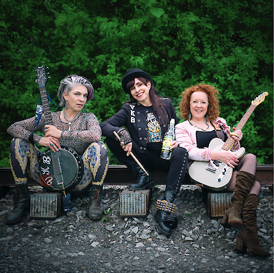 Das Bild zeigt eine Musikgruppe bestehend aus drei Personen, die auf Verstärkern oder Boxen sitzen vor einem grünen Hintergrund aus Pflanzen. Die Musiker halten verschiedene Instrumente - eine Banjo/Banjo-Gitarre links, ein Schlagzeug-Stick und eine Flasche in der Mitte, sowie eine E-Gitarre rechts - und tragen unterschiedliche stilvolle Outfits mit einem rockigen, alternativen Erscheinungsbild.