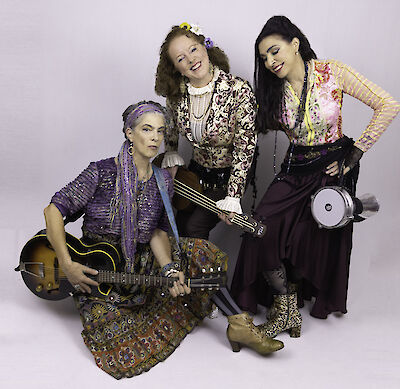 Das Bild zeigt eine Musikgruppe bestehend aus drei Personen in bunter, bohemischer Kleidung vor einem weißen Hintergrund. Eine Person sitzt mit einer Gitarre, während die anderen beiden stehen, eine mit einem Tamburin, alle tragen auffälligen Schmuck und farbenprächtige Outfits im Folk- oder Vintage-Stil.