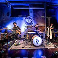 Das Bild zeigt eine Musikband, die in einem atmosphärisch blau beleuchteten Raum auftritt. Die Musiker spielen verschiedene Instrumente wie ein Saiteninstrument, Gitarre, Schlagzeug und Blasinstrumente auf einer kleinen Bühne mit einem dekorativen Emblem an der Wand im Hintergrund.