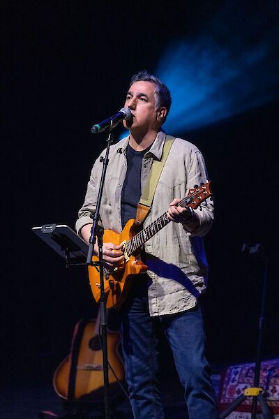 Das Bild zeigt einen Musiker während eines Live-Auftritts, der eine orange-gelbe E-Gitarre spielt und vor einem Mikrofon singt. Er trägt ein beiges Hemd über einem dunklen T-Shirt und steht auf einer Bühne mit dramatischer blauer Beleuchtung im Hintergrund.