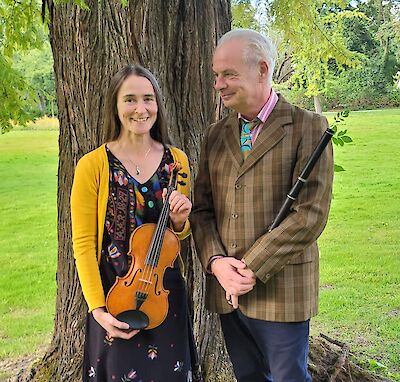 Das Bild zeigt ein Musikerduo bestehend aus einer Frau mit Violine in gelbem Cardigan und gemustertem Kleid sowie einem Mann mit Flöte in kariertem Anzug, die vor einem großen Baumstamm in einem Park stehen. Die beiden Musiker lächeln einander zu und befinden sich in einer grünen Parklandschaft mit Wiese und Bäumen im Hintergrund.