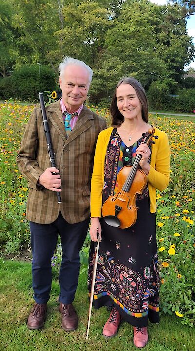 Das Bild zeigt zwei Musiker in einem Park mit bunten Blumen: Ein älterer Mann im karierten Anzug hält eine schwarze Flöte, während eine Frau in gelbem Cardigan und gemustertem Kleid eine Violine präsentiert. Die beiden stehen lächelnd auf einer Wiese vor einem üppigen Blumenbeet mit gelben Blüten und grünen Bäumen im Hintergrund.