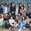Das Bild zeigt eine fröhliche Gruppe von etwa 20 jungen Menschen, die vor einer blauen Holzwand posieren und dabei lächeln oder lachen. Die Jugendlichen und jungen Erwachsenen sind in legerer, bunter Freizeitkleidung gekleidet und arrangieren sich in zwei Reihen - einige stehen im Hintergrund, andere sitzen oder hocken im Vordergrund auf dem Pflasterboden.