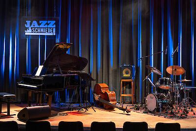 Das Bild zeigt eine Konzertbühne mit verschiedenen Musikinstrumenten, darunter ein schwarzer Flügel, ein Kontrabass, ein Schlagzeug und weitere Equipment, alles atmosphärisch mit blauem Licht beleuchtet. Im Hintergrund ist ein Schild mit der Aufschrift "JAZZ SCHMIEDE" zu sehen, was darauf hinweist, dass es sich um eine Jazz-Spielstätte handelt.