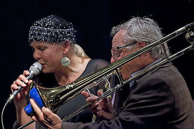 Eine Jazzsängerin mit glitzernder Kopfbedeckung singt in ein Mikrofon, während neben ihr ein älterer Posaunist mit Brille sein Instrument spielt. Die beiden Musiker performen gemeinsam auf einer Bühne vor dunklem Hintergrund in einer intimen Konzertszene.