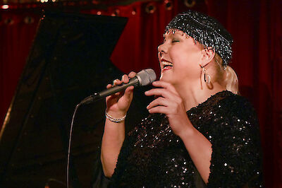 Eine Jazzsängerin mit glitzernder Kopfbedeckung und schwarzem Paillettenkleid singt leidenschaftlich in ein Mikrofon auf einer Bühne. Im Hintergrund sind warme rötliche Bühnenbeleuchtung und ein Flügel zu erkennen, was eine typische Jazz-Club-Atmosphäre schafft.