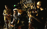 Eine Band bestehend aus vier Musikern tritt in einem Stadtgarten auf, erkennbar an der Beleuchtung und dem Schriftzug "STADTGARTEN" im Hintergrund. Die Gruppe umfasst eine Sängerin mit Mikrofon in der Mitte sowie Musiker mit Gitarre, Bass und weiteren Instrumenten in stimmungsvollem Bühnenlicht.