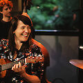 Eine Frau mit dunklen Haaren lacht herzlich, während sie eine Ukulele spielt und vor einem Mikrofon sitzt. Im Hintergrund sind weitere Musiker zu sehen, darunter ein Bassist, was auf eine Live-Aufnahme oder ein Konzert in entspannter Atmosphäre hindeutet.