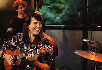 Eine Frau mit dunklen Haaren lacht herzlich, während sie eine Ukulele spielt und vor einem Mikrofon sitzt. Im Hintergrund sind weitere Musiker zu sehen, darunter ein Bassist, was auf eine Live-Aufnahme oder ein Konzert in entspannter Atmosphäre hindeutet.