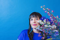 Eine Frau mit dunklen Haaren und Pony trägt ein blaues Oberteil und posiert vor einem leuchtend blauen Hintergrund. Sie hält einen bunten Blumenstrauß mit violetten, weißen und rosa Blüten, der harmonisch mit dem farbenfrohen Setting komponiert ist.