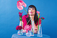 Eine Frau mit dunklen Haaren und Pony sitzt vor einem blauen Hintergrund an einem Tisch mit verschiedenen Glasvasen voller Blumen in Rosa- und Pinktönen. Sie trägt ein pinkes Oberteil, stützt ihr Kinn nachdenklich auf die Hand und ist von bunten Blüten wie Anthurien, Rosen und Chrysanthemen umgeben.