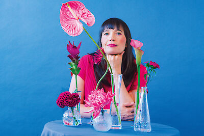 Eine Frau mit dunklen Haaren und Pony sitzt vor einem blauen Hintergrund an einem Tisch mit verschiedenen Glasvasen voller Blumen in Rosa- und Pinktönen. Sie trägt ein pinkes Oberteil, stützt ihr Kinn nachdenklich auf die Hand und ist von bunten Blüten wie Anthurien, Rosen und Chrysanthemen umgeben.