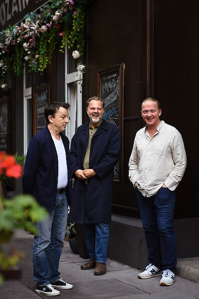 Drei Männer stehen vor einem Restaurant. Sie lächeln. Über dem Eingang sind Blumen. An der Tür hängt eine Tafel. Die Männer tragen Jeans und Hemden. Sie machen ein Foto zusammen. Sie stehen auf dem Gehweg.