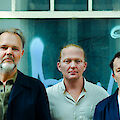 Three men pose for a group photo in front of a turquoise-coloured wall with large windows. The man on the left is wearing a dark jacket and has a grey beard, the man in the middle is wearing a light-coloured shirt and the man on the right is wearing a white T-shirt under a dark jacket.