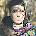 The portrait shows a person with short dark hair and a nose piercing, wearing a colourful patterned scarf and dark clothing. The photo was taken outdoors in natural light, with shadows from trees or branches falling on the face and the background blurred.