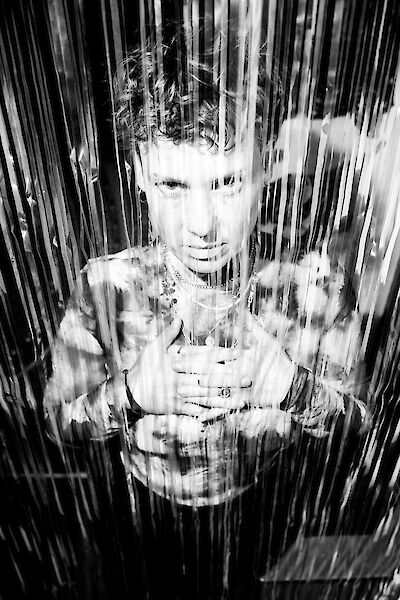 The black and white photo shows a person with curly hair and a necklace standing behind a curtain of vertical ribbons or stripes. The person looks through the hanging elements into the camera, creating an interesting visual effect with plays of light and shadow.