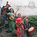 Four musicians in colourful, traditional clothing pose with their instruments on an outdoor staircase in front of a white wall. The group holds various stringed instruments and a drum and is surrounded by green plants in terracotta pots.