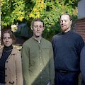 The picture shows four young musicians from the Simon Kuhn Quartet posing for a group photo in front of a brick wall with green trees in the background. From left to right, a woman in a beige coat, two men in a green shirt and a grey jumper respectively and another man in a dark shirt can be seen.