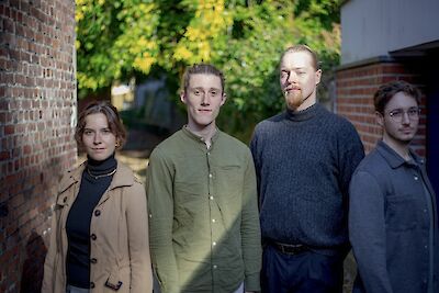 Das Bild zeigt vier junge Musiker. Sie sind vom Simon Kuhn Quartett. Sie machen ein Foto zusammen. Hinter ihnen ist eine Wand aus Steinen. Es gibt auch grüne Bäume im Hintergrund. Links steht eine Frau. Sie trägt einen beigen Mantel. Dann stehen zwei Männer da. Einer trägt ein grünes Hemd. Der andere trägt einen grauen Pullover. Rechts steht noch ein Mann. Er trägt ein dunkles Hemd.
