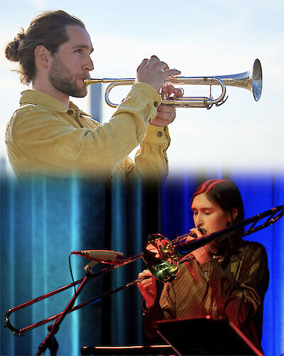 Das Bild zeigt zwei Musiker, die Blechblasinstrumente spielen - im oberen Bildbereich ein Mann mit längeren Haaren und Bart in gelblicher Jacke, der Trompete spielt, und im unteren Bereich eine Person in dunkler Kleidung, die Posaune spielt. Beide befinden sich offensichtlich bei einer Live-Aufführung oder einem Konzert, wobei im Hintergrund bläuliche Beleuchtung und Bühnentechnik zu sehen sind.