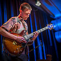 The picture shows a jazz guitarist during a live performance, concentrating on his brown and gold hollow-body guitar. The scene is bathed in atmospheric blue stage lighting, with other musicians and stage equipment recognisable in the background.