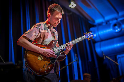 Das Bild zeigt einen Mann. Er spielt Jazz-Gitarre. Er tritt live auf. Er spielt konzentriert. Seine Gitarre ist braun und golden. Die Gitarre ist hohl. Die Bühne hat blaues Licht. Im Hintergrund sind andere Musiker. Da ist auch Technik für die Bühne.