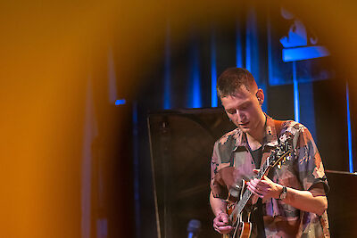 Das Bild zeigt einen jungen Mann. Er spielt Gitarre. Er trägt ein Hemd mit Muster. Er ist bei einem Konzert. Er spielt eine E-Gitarre. Er ist sehr konzentriert. Die Bühne hat warmes Licht. Das Licht ist orange-gelb und blau. Im Hintergrund sind andere Musiker. Da ist auch Technik für die Bühne.