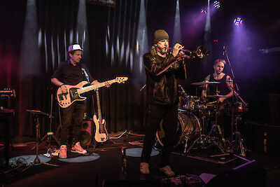 Eine Jazz-Band spielt auf der Bühne. Die Band hat drei Musiker. In der Mitte steht ein Trompeter. Er trägt eine Mütze und eine Lederjacke. Links spielt ein Bassist. Er trägt eine Basecap. Rechts sitzt ein Schlagzeuger. Violettes Licht macht die Bühne schön. Es ist ein Live-Konzert.