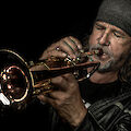A man with a grey woolly hat and long hair concentrates on playing the trumpet against a black background. The golden instrument gleams in the warm light, while the musician is completely absorbed in his playing with his eyes closed.