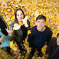 Four young people lie smiling in a large pile of colourful autumn leaves and playfully throw the yellow and brown leaves into the air. The group consists of three women and a man with glasses, all looking happily into the camera and enjoying the autumnal moment.
