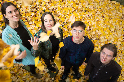 Four young people lie smiling in a large pile of colourful autumn leaves and playfully throw the yellow and brown leaves into the air. The group consists of three women and a man with glasses, all looking happily into the camera and enjoying the autumnal moment.