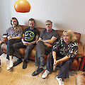 Four people sit relaxed next to each other on a brown leather sofa in a modern room with an orange and yellow hanging lamp. The group consists of three men and one woman, all dressed casually and looking at the camera in a friendly manner.
