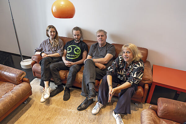 Four people sit relaxed next to each other on a brown leather sofa in a modern room with an orange and yellow hanging lamp. The group consists of three men and one woman, all dressed casually and looking at the camera in a friendly manner.