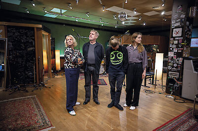 4 Menschen stehen in einem Musik-Studio. Es sind 2 Frauen und 2 Männer. Sie schauen nach oben. Im Studio gibt es viele Geräte für Musik. Es gibt Mikrofone und Verstärker. An der Decke hängt eine Diskokugel. Der Boden ist aus Holz.