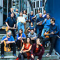 The picture shows a large group of around 20 musicians with various instruments such as saxophone, trombone, bass and guitar, posing on a stone staircase in front of a modern glass building. The musicians wear predominantly blue clothing and are arranged in several rows on the steps, with two women in striking red and burgundy-coloured outfits sitting in the foreground.