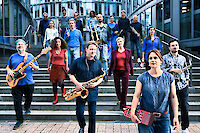 Das Bild zeigt etwa 20 Musiker. Sie haben verschiedene Instrumente. Zum Beispiel Saxophon, Posaune und Bass. Das sind Jazz-Instrumente. Die Musiker stehen auf einer Steintreppe. Hinter ihnen ist ein modernes Glasgebäude. Die meisten Musiker tragen blaue Kleidung. Einige haben rote Akzente. Sie stehen in mehreren Reihen auf den Stufen.