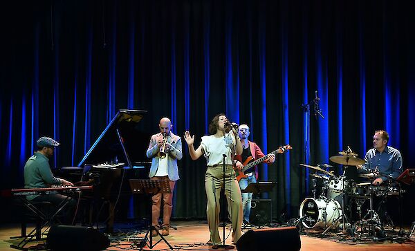 A five-piece band performs on stage with a singer in the centre standing in front of a microphone, accompanied by musicians on piano, trumpet, bass and drums. The scene is illuminated by dramatic blue strips of light in the background, creating an atmospheric concert atmosphere.