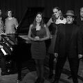 The black and white photo shows a group of eight musicians of different ages, elegantly dressed and posing around a grand piano. On the left, a pianist sits at the open instrument, while the other band members are arranged in dark, stylish clothing for a professional group photo.