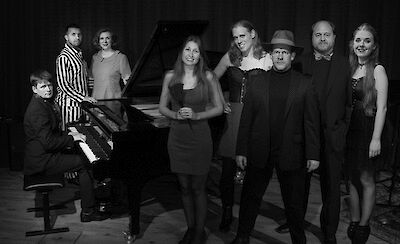 Das ist ein Schwarz-Weiß-Foto. Es zeigt acht Musiker. Die Musiker sind alt und jung. Sie tragen schöne Kleidung. Sie stehen um einen Flügel. Links sitzt ein Pianist. Er spielt am Flügel. Die anderen Musiker stehen daneben. Sie tragen dunkle Kleidung. Das ist ein Gruppen-Foto.