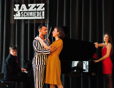 Das Bild zeigt eine Theater-Szene. Die Szene ist in der Jazzschmiede. Da sind vier Personen. Links sitzt ein Mann am Klavier. In der Mitte stehen zwei Personen. Das ist ein Mann und eine Frau. Der Mann trägt einen Anzug mit Streifen. Die Frau trägt ein gelbes Kleid. Sie umarmen sich. Rechts steht eine Frau. Sie trägt ein rotes Kleid. Sie steht am Flügel. Das sieht aus wie ein Musical. Die Stimmung ist romantisch.