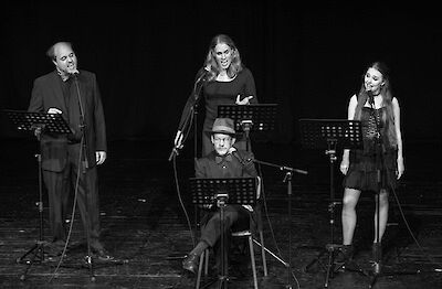 Das Foto ist schwarz-weiß. Es zeigt vier Musiker auf der Bühne. Drei Personen stehen am Mikrofon. Sie singen. Eine Person sitzt am Klavier. Sie spielt Musik. Um die Musiker stehen Notenständer. Es gibt auch Bühnentechnik. Die Musiker geben ein Konzert.