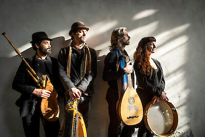 The picture shows four musicians from the Toasaves ensemble in dark clothing holding various traditional instruments such as bagpipes, lute and frame drum. The group stands in front of a light-coloured wall with dramatic strips of light falling through windows, creating a moody atmosphere.