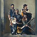 The picture shows four musicians from the ensemble Toasaves posing with various traditional instruments such as lutes, flutes and other historical musical instruments. The group is arranged in a rustic room with weathered walls and wears timeless, earthy clothing that matches their concept of traditional Flemish music.