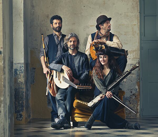 The picture shows four musicians from the ensemble Toasaves posing with various traditional instruments such as lutes, flutes and other historical musical instruments. The group is arranged in a rustic room with weathered walls and wears timeless, earthy clothing that matches their concept of traditional Flemish music.