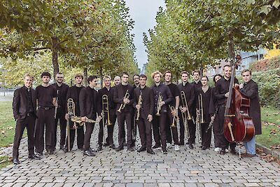 Das Bild zeigt eine große Jazzband mit etwa 20 Musikern in schwarzer Kleidung, die mit ihren Instrumenten wie Trompeten, Posaunen, Saxophonen und einem Kontrabass in einem Park posieren. Die Musiker stehen auf einem gepflasterten Weg zwischen grünen Bäumen, im Hintergrund sind moderne Gebäude zu sehen.