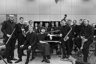 Das schwarz-weiße Foto zeigt eine große Jazzband mit etwa 18 Musikern, die in einem modernen Proberaum um ein Klavier gruppiert sind. Die Musiker halten verschiedene Instrumente wie Trompeten, Posaunen und Saxophone und sind alle in dunkler Kleidung für ein Gruppenfoto aufgestellt.