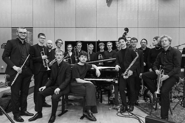 The black and white photo shows a large jazz band of around 18 musicians grouped around a piano in a modern rehearsal room. The musicians are holding various instruments such as trumpets, trombones and saxophones and are all dressed in dark-coloured clothing for a group photo.