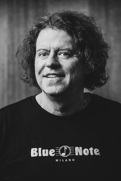 The black and white portrait photo shows a smiling middle-aged man with curly, dark hair and a T-shirt with "Blue Note Milano" printed on it. The musician looks friendly into the camera, while a room with columns or pillars can be recognised blurred in the background.