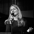 The black and white photo shows a young woman with long blonde hair singing into a microphone and smiling. She is wearing a dark-coloured top and is obviously performing live on stage.